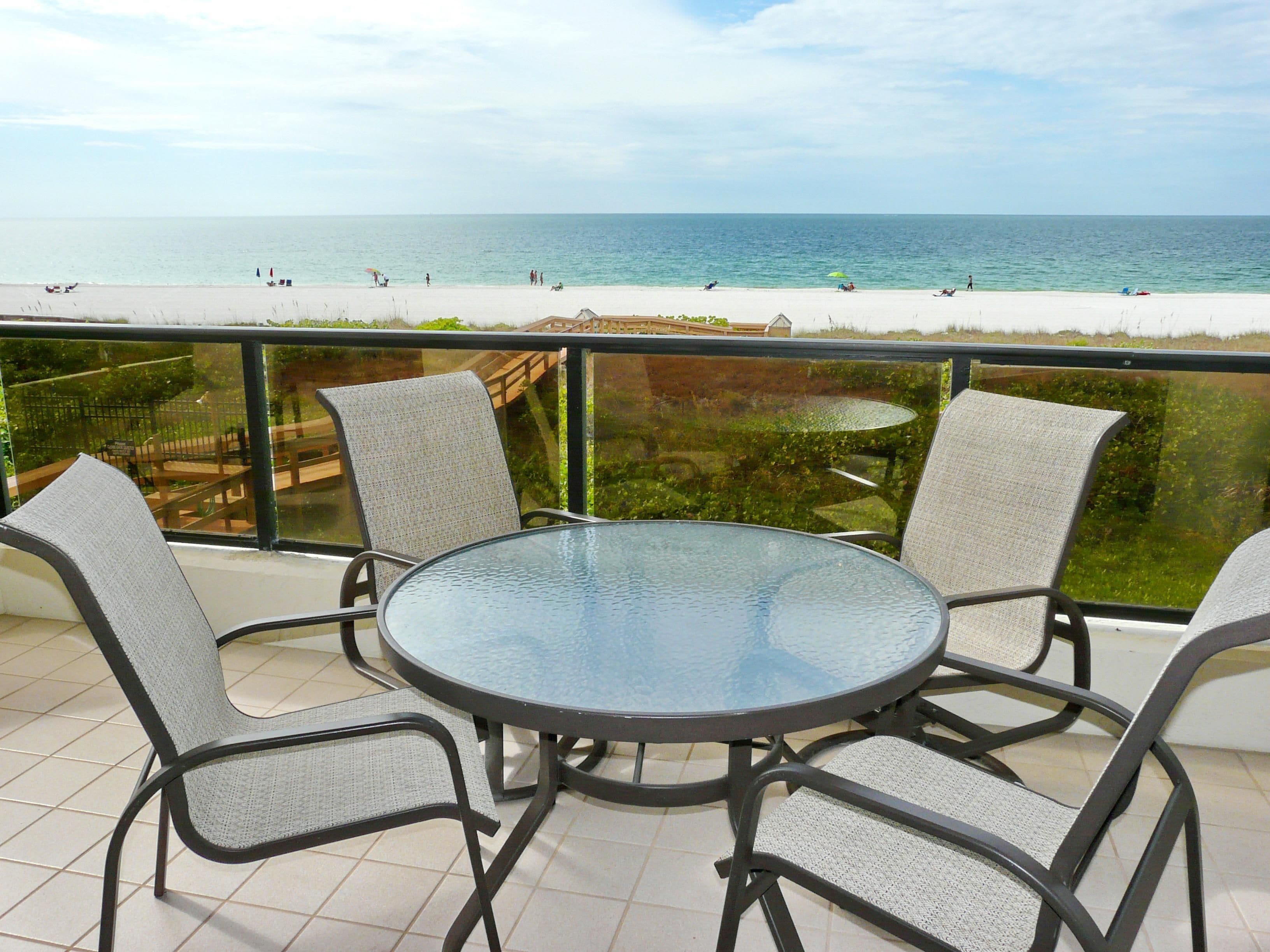 A round glass table with four chairs on a balcony overlooking a beach.