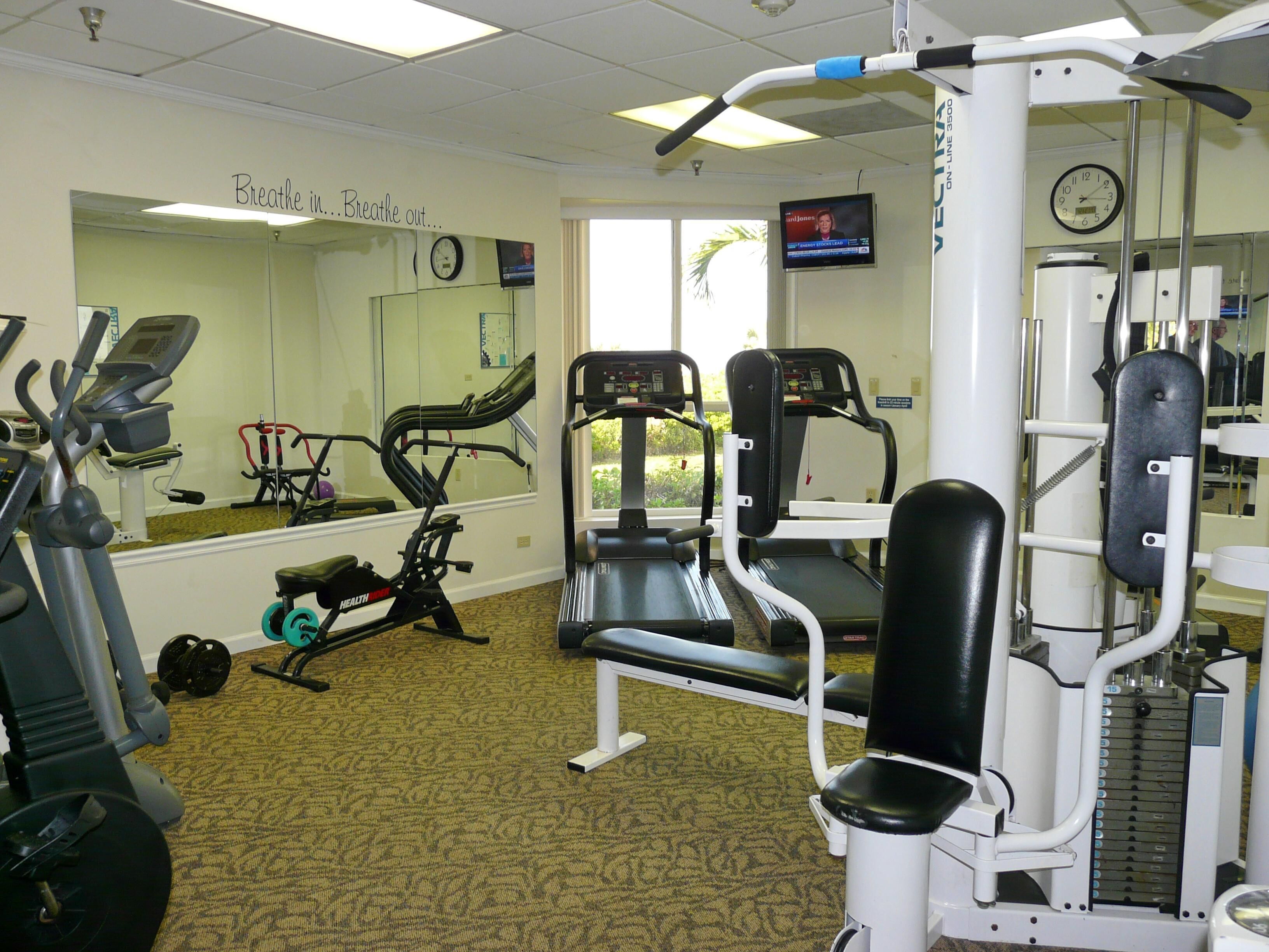 A gym with exercise equipment, a mirror, a clock, and a TV screen.