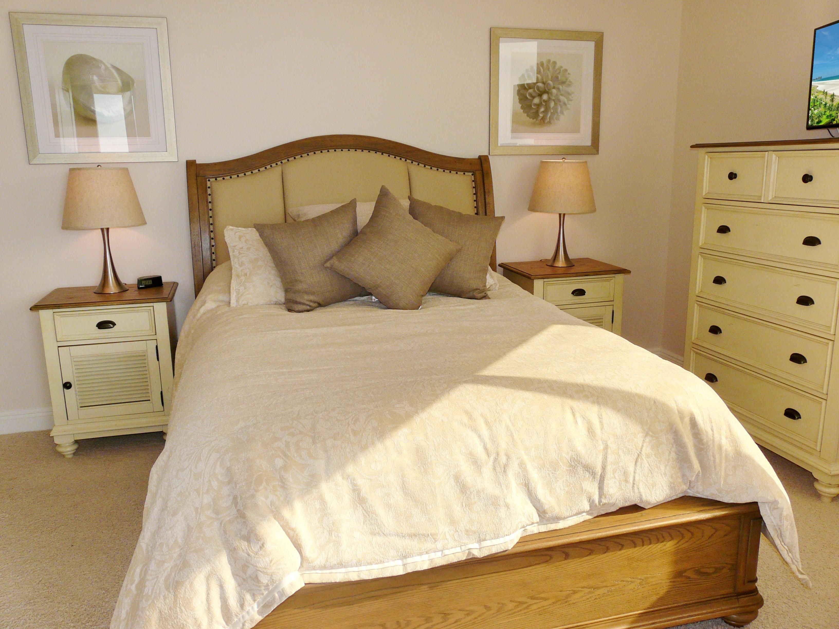 Una cama bien hecha con tablero de madera, dos mesitas de noche con lámparas y un aparador con varios cajones.