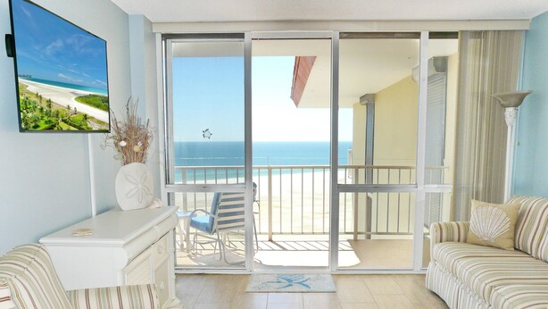 Un salon spacieux donnant sur la plage grâce à une porte-fenêtre coulissante, avec un canapé blanc, un fauteuil à rayures et une petite table agrémentée d’objets décoratifs.