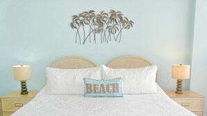 Un lit recouvert d’une couvre-lit blanche, deux tables de chevet avec des lampes, une tête de lit et une décoration murale représentant des palmiers.