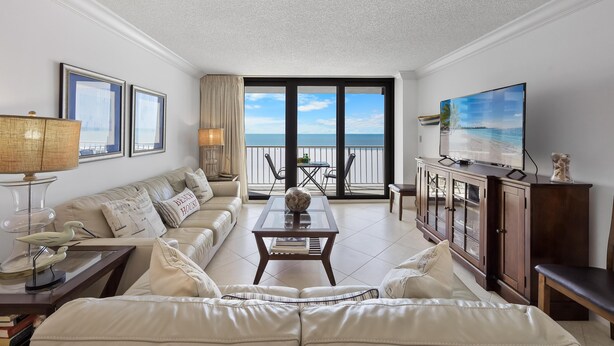 A living room with a sofa, coffee table, and TV, with a view of the ocean through large windows. - Eye-catching Beachfront Condo With Pool and Nice Views (Marco Island)