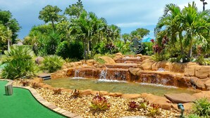 A landscaped area with a water feature, including a cascading waterfall and a pond, surrounded by various tropical plants and trees. - Inviting Beachfront Condo With Pool, spa and Views (Marco Island)