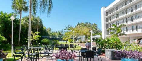 An outdoor patio area with tables and chairs, surrounded by greenery and a white building in the background.