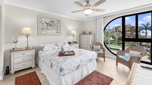 A bedroom with a bed, nightstands, a dresser, a chair, and a large window offering a view of palm trees and buildings. - Stylish Condo Across From South Beach (Marco Island)