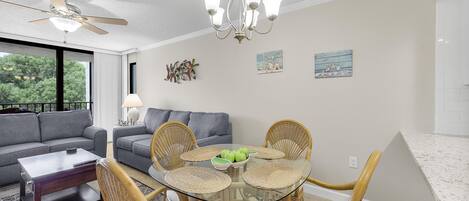 A modern living room with a glass dining table surrounded by wicker chairs, a gray sofa, and a ceiling fan.