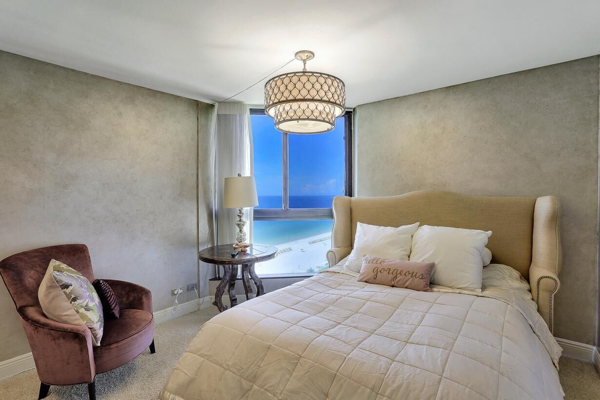 A bedroom with a large bed, a chair, a table, and a view of the sea through a window.