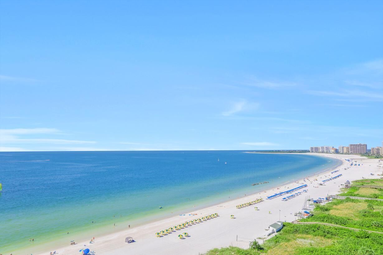 A beach with rows of umbrellas and beach chairs, a clear blue sky, and a calm sea.