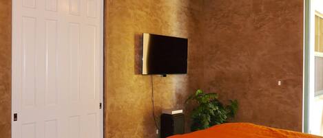 Un dormitorio con una puerta blanca, televisor montado, una planta y una colcha naranja.