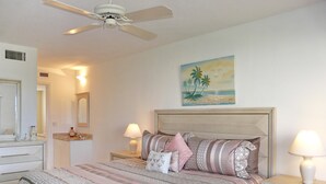 A bedroom with a bed, nightstands, a ceiling fan, a painting of a beach, and a mirror. - Coastal Style Condo With Pool, spa and Steps to Beach (Marco Island)