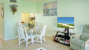 A dining area with a glass table and white chairs, a kitchen in the background, a painting on the wall, and a TV on a stand. - Coastal Style Condo With Pool, spa and Steps to Beach (Marco Island)