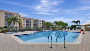 A swimming pool with a tiled deck, surrounded by a fence and palm trees, with a building in the background. - Coastal Style Condo With Pool, spa and Steps to Beach (Marco Island)