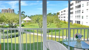 A balcony with a glass table and wicker chair, overlooking a well-maintained lawn and a small gazebo. - Coastal Style Condo With Pool, spa and Steps to Beach (Marco Island)