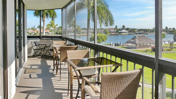 A balcony with wicker furniture, a glass table, and a view of a lake and houses.