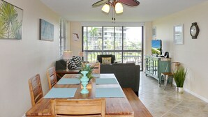 A dining area with a wooden table, chairs, and a ceiling fan. There is a window with a view of buildings and greenery outside.