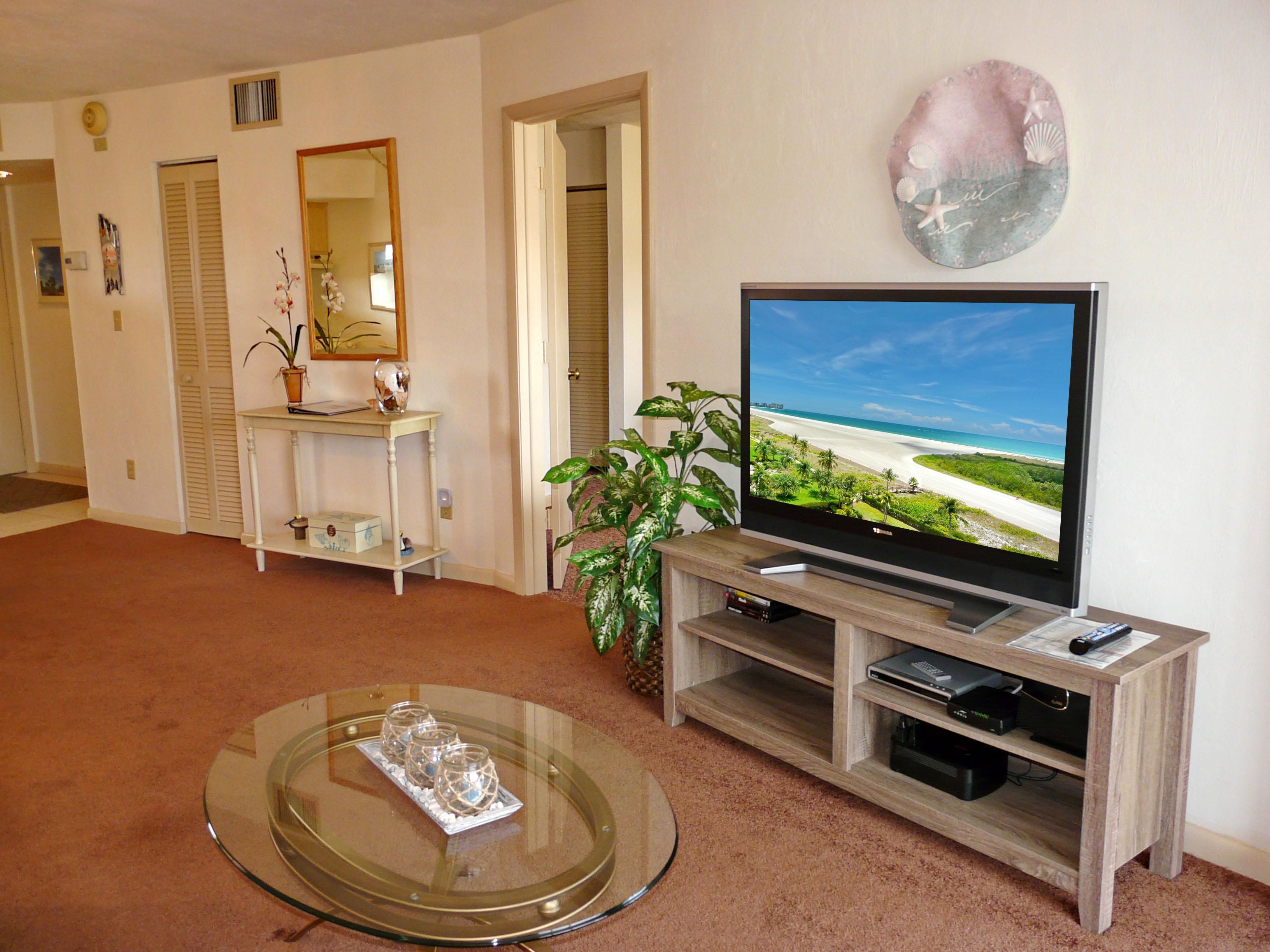 A living room with a flat-screen TV on a wooden stand, a glass coffee table with decorative items, a potted plant, and a beach scene on the TV.