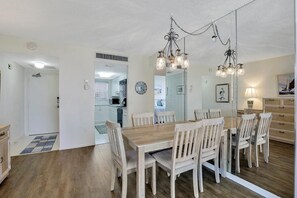 Uma sala de jantar com uma mesa e cadeiras de madeira, um lustre e uma cozinha ao fundo.