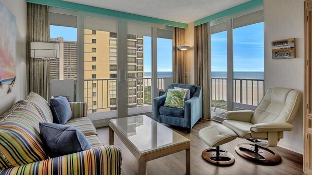 A modern hotel room with a balcony, a striped sofa, a recliner, a glass coffee table, and a view of the ocean.