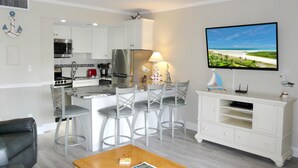 A compact living space with a kitchenette, a dining area with bar stools, a flat-screen TV mounted on the wall, and a wooden coffee table. - Beach Club 212 (Marco Island)