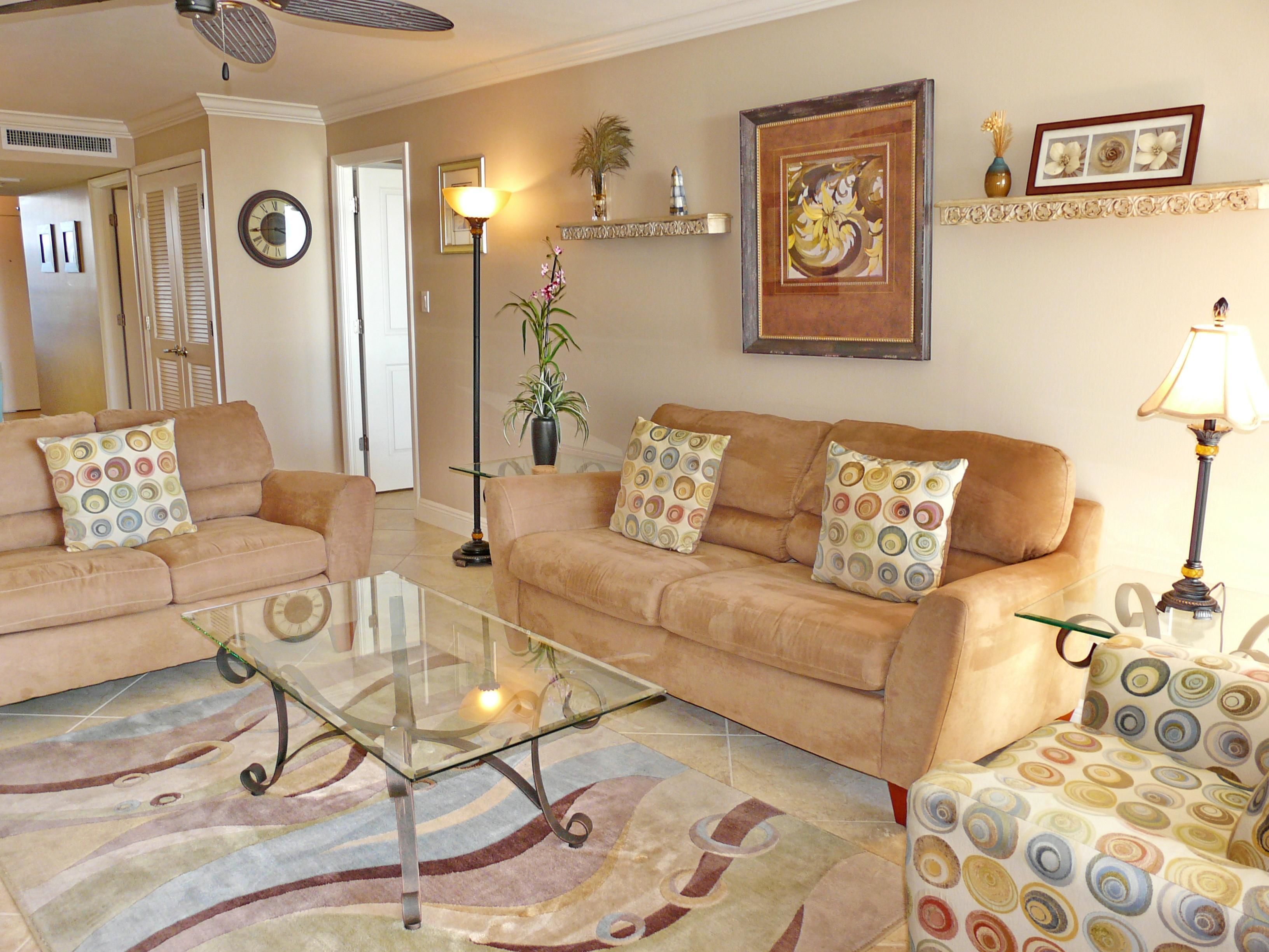 Una sala de estar con un sofá color beige, una mesa de centro de vidrio y cojines estampados. Hay una planta en maceta, una obra de arte enmarcada y una lámpara en una mesita auxiliar.