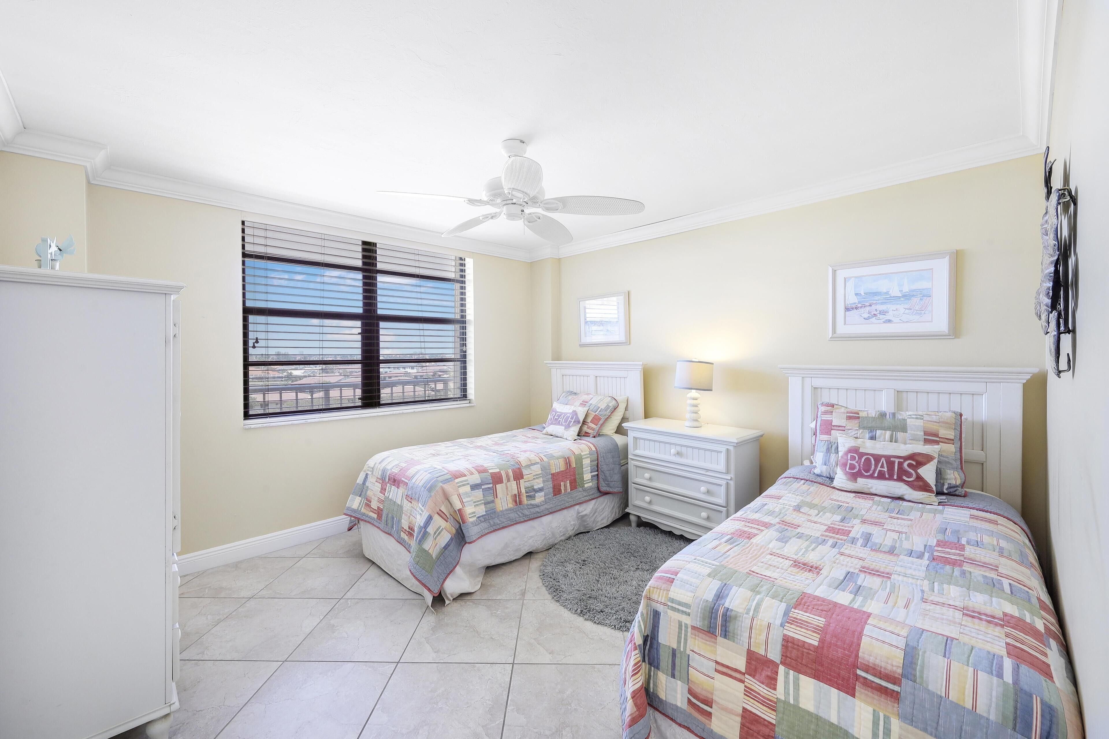 A bedroom with two beds, a ceiling fan, a window with blinds, a nightstand with a lamp, and a framed picture on the wall.