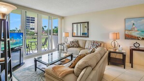 A living room with a sofa, coffee table, and TV, overlooking a balcony with a view of a building and palm trees. - Cozy Beachfront Condo With Pool and Tennis Courts (Marco Island)