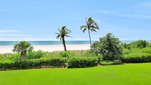 A lush green lawn, palm trees, and a beach with clear skies. - Cozy Beachfront Condo With Pool and Tennis Courts (Marco Island)