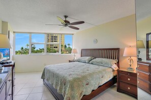 A bedroom with a bed, a ceiling fan, a nightstand, a dresser, and a window with a view of palm trees and buildings. - Cozy Beachfront Condo With Pool and Tennis Courts (Marco Island)