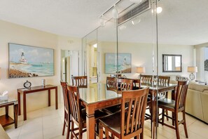 A dining area with a glass-top table, wooden chairs, and a painting of a lighthouse. - Cozy Beachfront Condo With Pool and Tennis Courts (Marco Island)