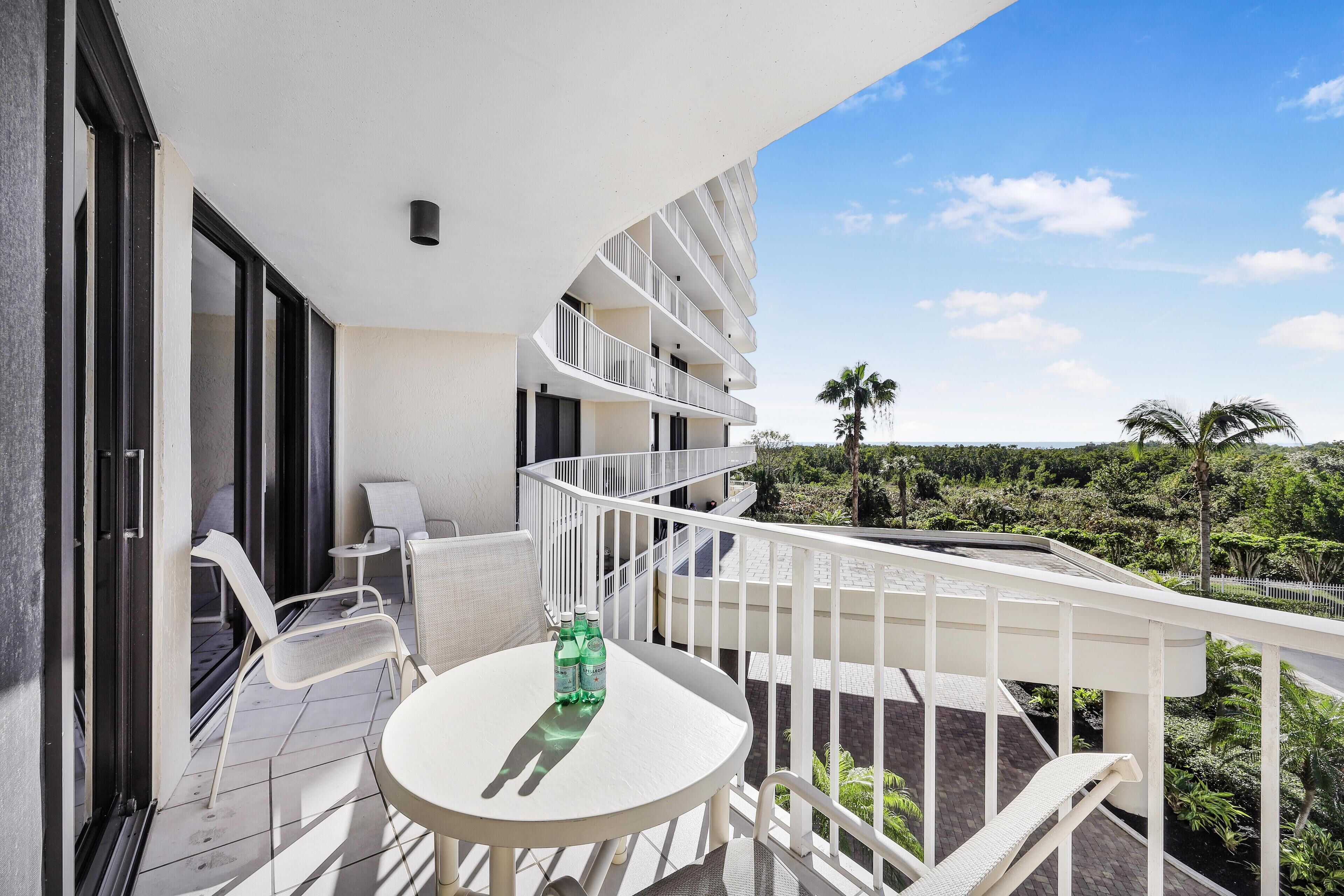 Ein Balkon mit einem weißen Tisch und Stühlen, zwei Wasserflaschen und Blick auf eine angelegte Grünfläche mit Palmen.