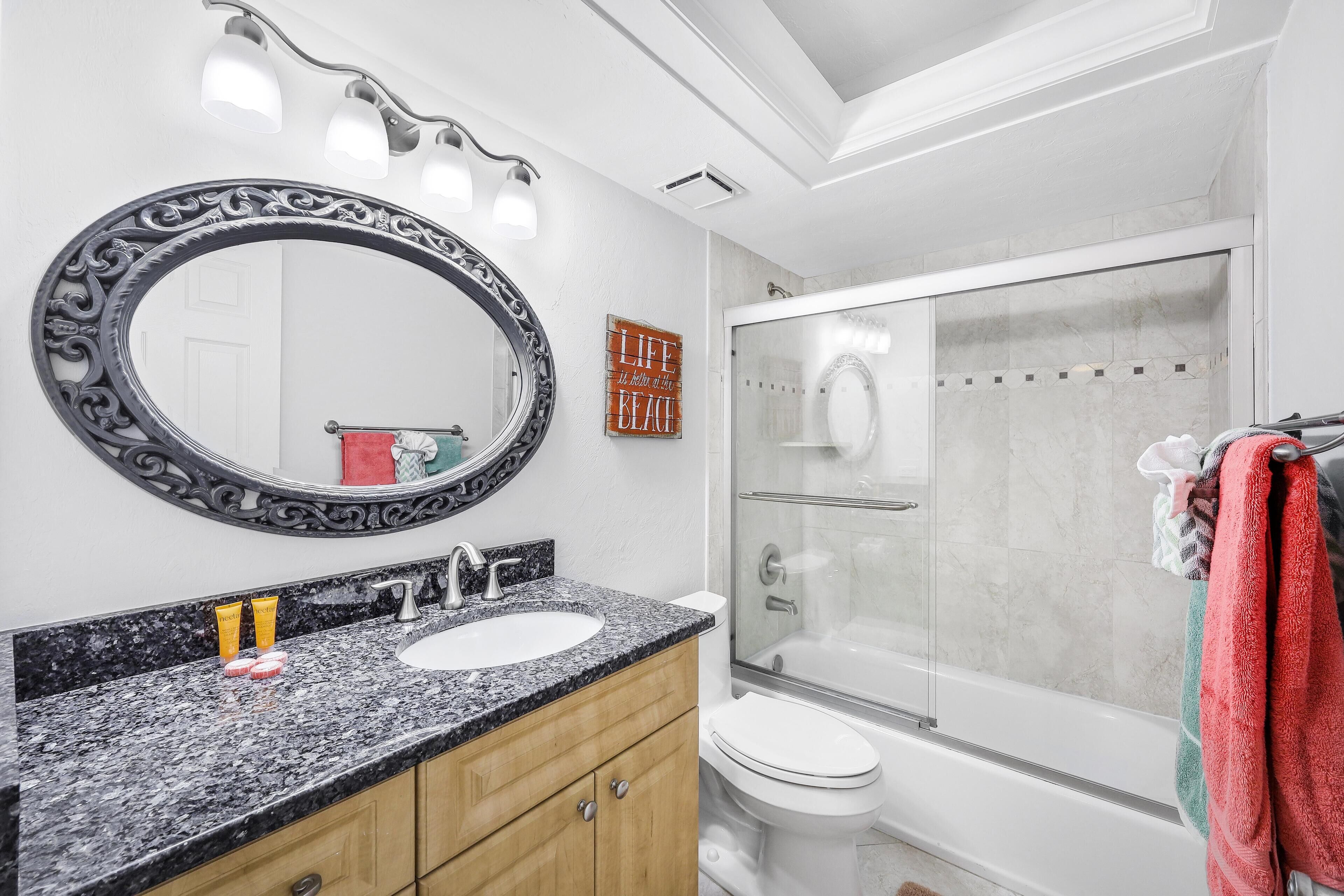 A bathroom with a granite countertop, a large mirror, a toilet, and a glass-enclosed shower.