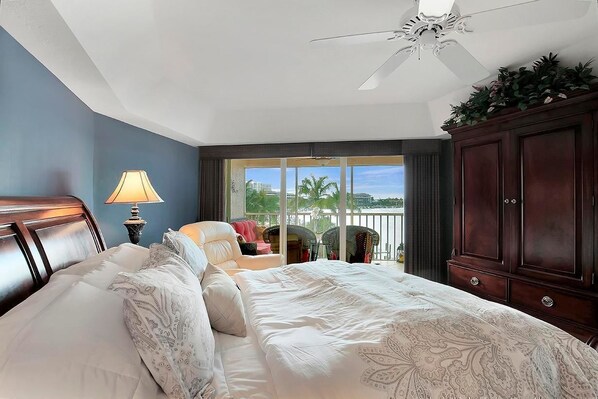 A hotel room with a large bed, a ceiling fan, a view of palm trees, and a balcony.