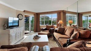 A spacious living room with a large flat-screen TV, comfortable seating, and a balcony with a view.