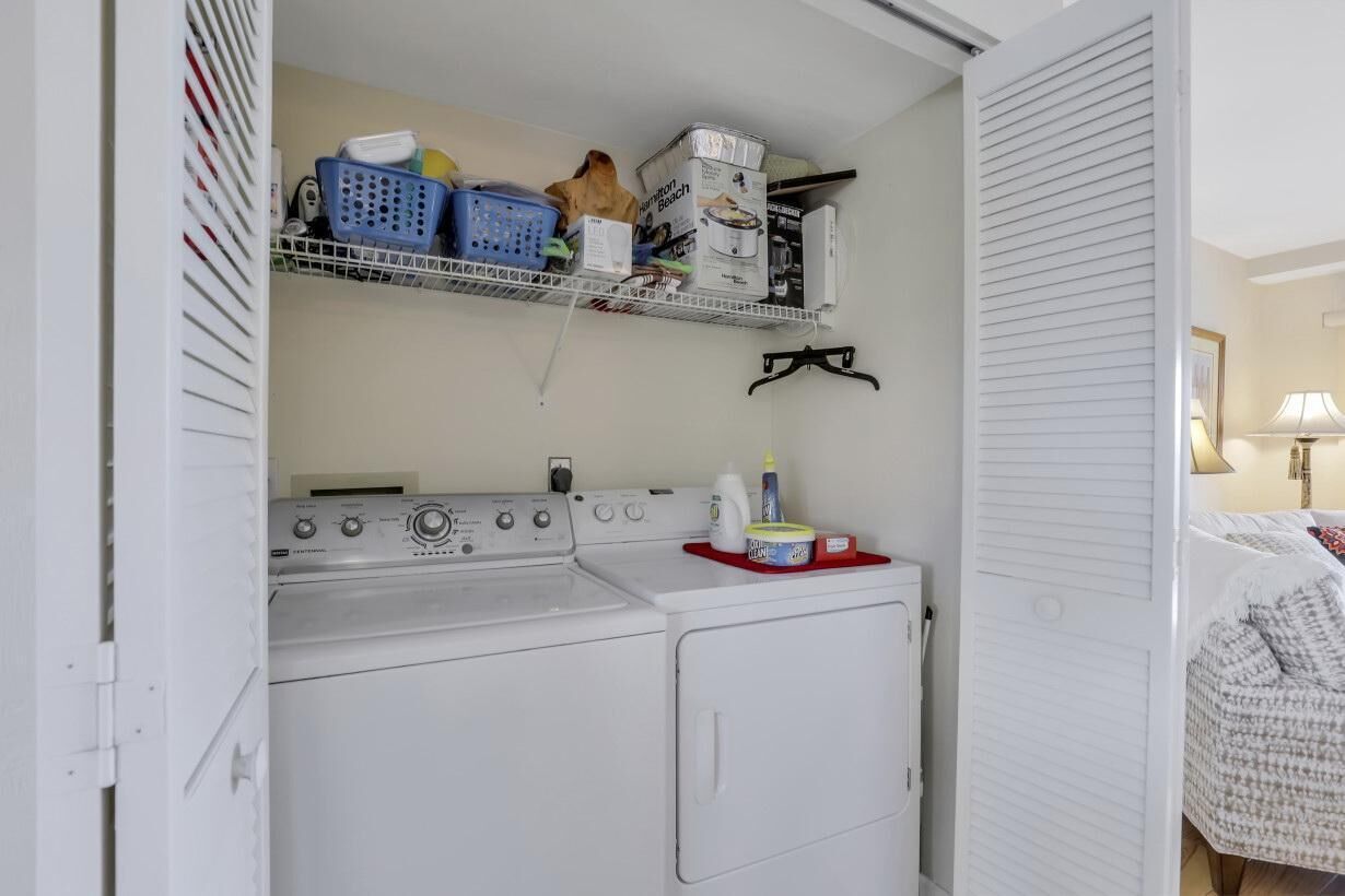 A laundry area with a washing machine and dryer, a shelf with baskets and cleaning supplies, and a bed with a patterned blanket.