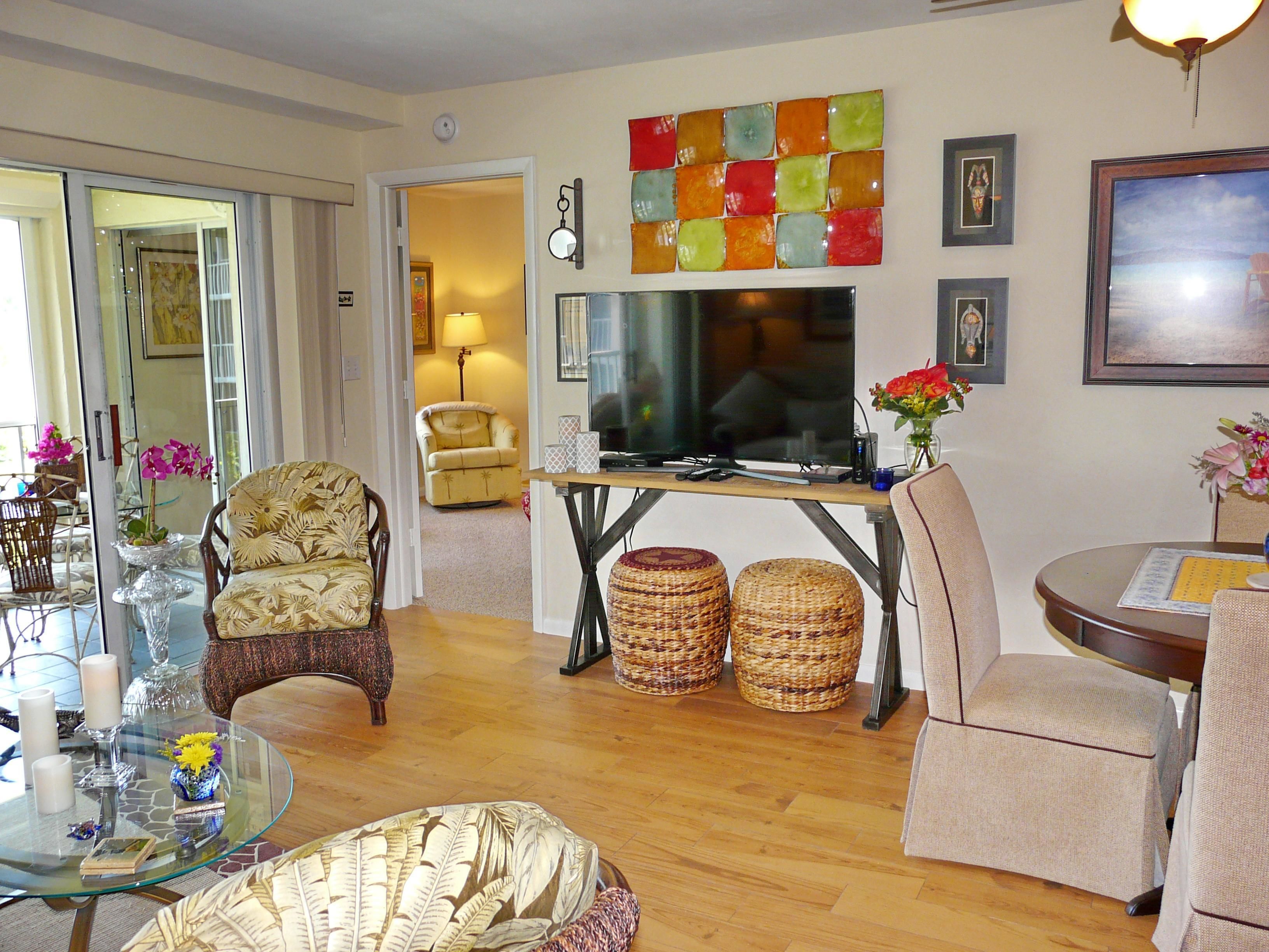 Un salon avec une table basse en verre, deux poufs en osier, un téléviseur et un fauteuil à motifs floraux.