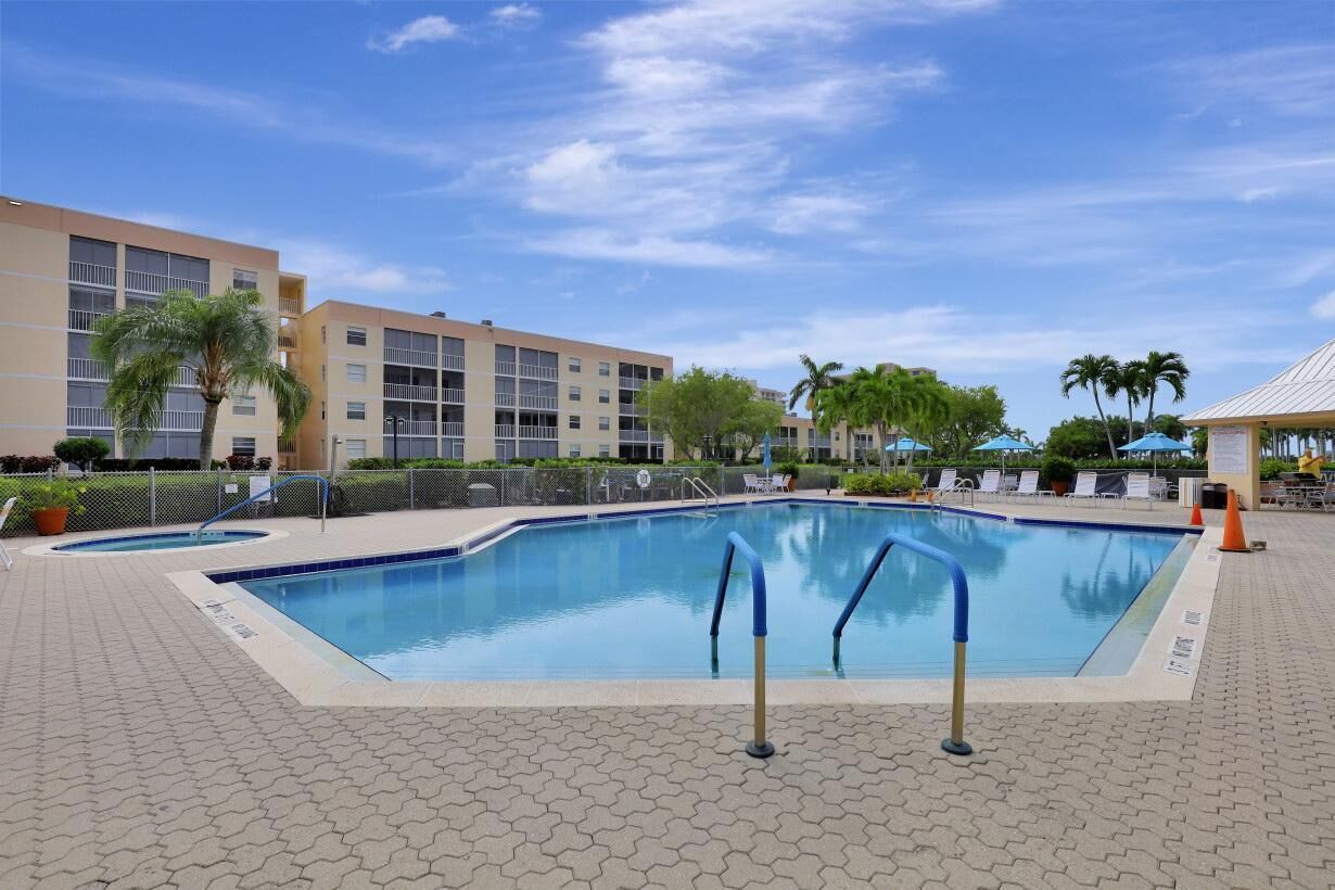 A swimming pool with a tiled deck, surrounded by a fence and palm trees, with a building in the background.