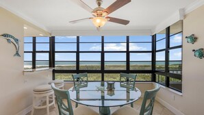 A dining area with a round glass table, four chairs, a ceiling fan, and large windows offering a view of the outdoors. - Beachfront Condo With Pool and Dazzling Ocean View (Marco Island)