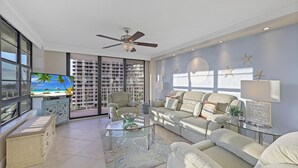A spacious living room with a large TV, comfortable seating, a ceiling fan, and a view of a high-rise building through large windows. - Beachfront Condo With Pool and Dazzling Ocean View (Marco Island)