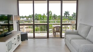 A living room with a sofa, TV, and a balcony with a view of palm trees. - Inviting Beachfront Condo With Large Balcony and Pool (Marco Island)