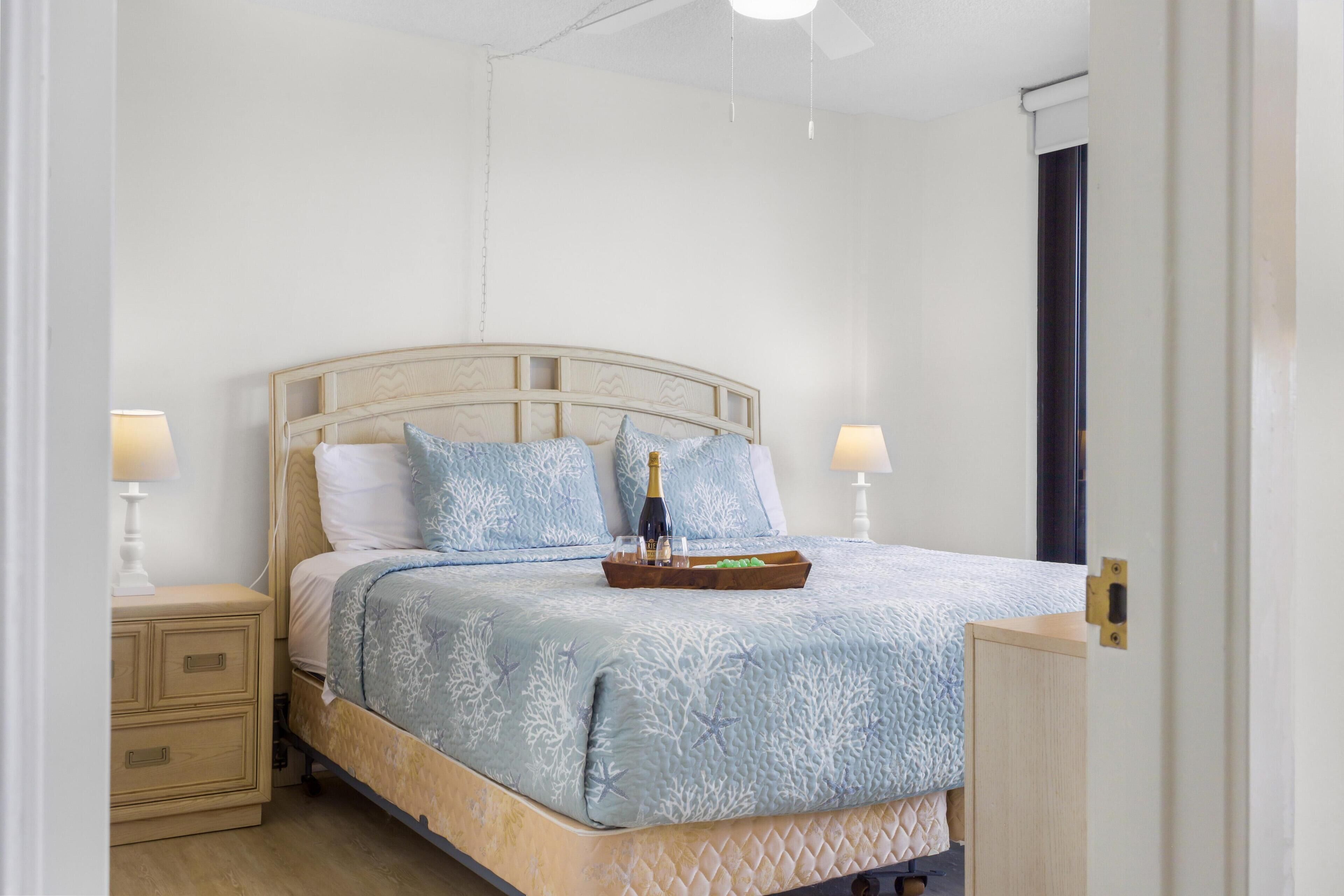 Un lit bien fait, recouvert d’une couvre-lit à motifs bleus, avec deux lampes de chevet et un plateau sur lequel se trouvent une bouteille et des verres.
