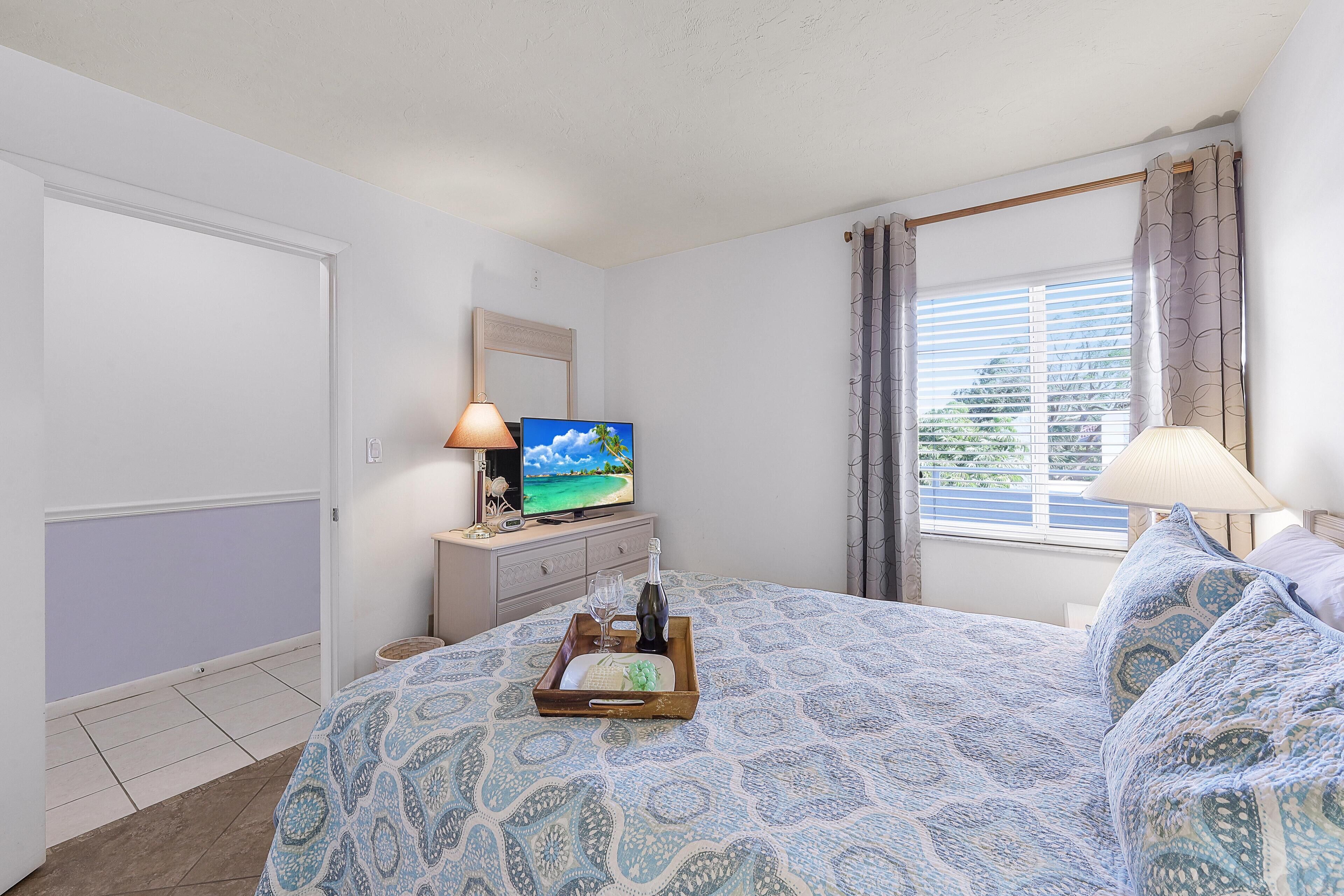Ein Schlafzimmer mit Bett, Fernseher, Kommode und einem Fenster mit Vorhängen.