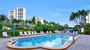 A swimming pool with lounge chairs and umbrellas, surrounded by palm trees and buildings.
