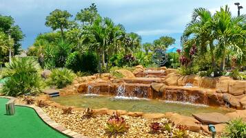 A landscaped area with a water feature, including a cascading waterfall and a pond, surrounded by various tropical plants and trees.