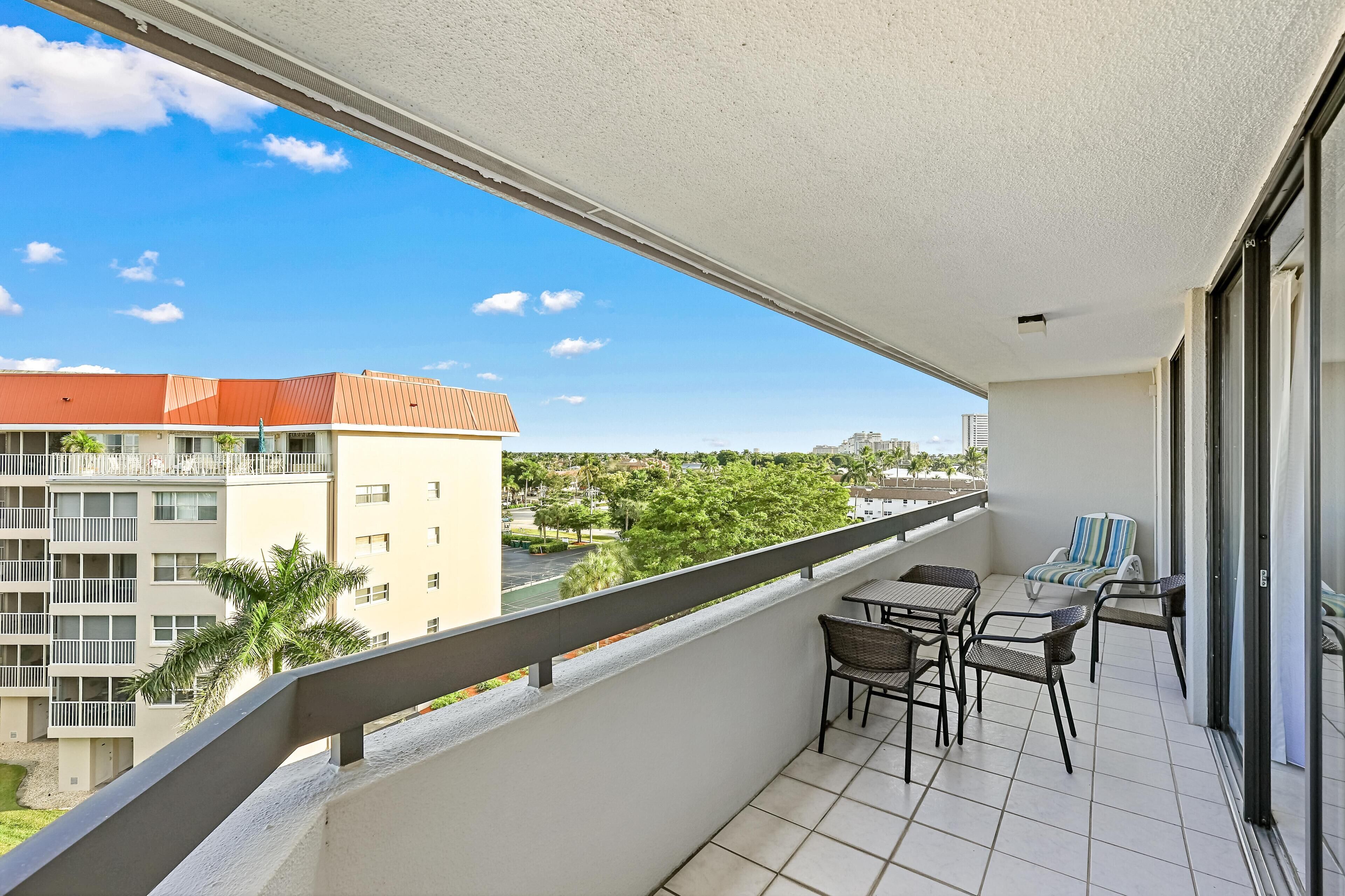 A balcony with a view of a cityscape, including buildings and greenery. There are two wicker chairs and a small table.