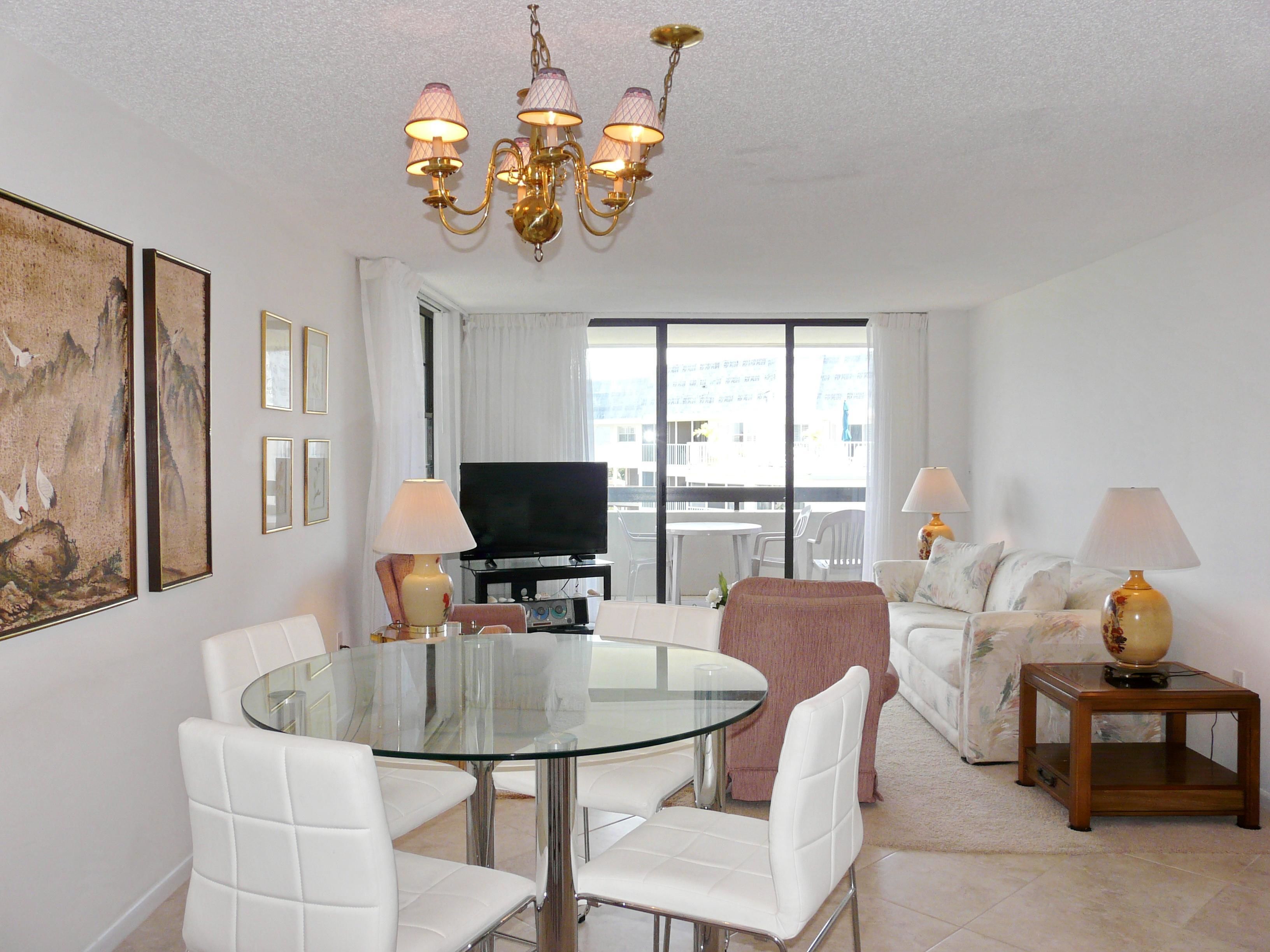 A modern living room with a glass dining table, white chairs, a sofa, a TV, and a chandelier.