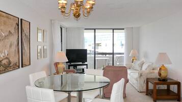 A modern living room with a glass dining table, white chairs, a sofa, a TV, and a chandelier.