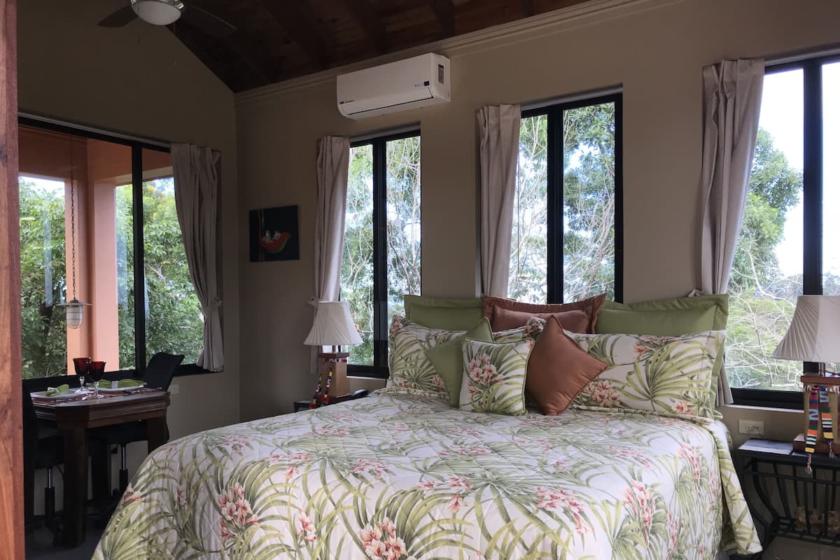 A bedroom with a bed, a desk, and a chair. There are windows with curtains, an air conditioning unit, and a ceiling fan.