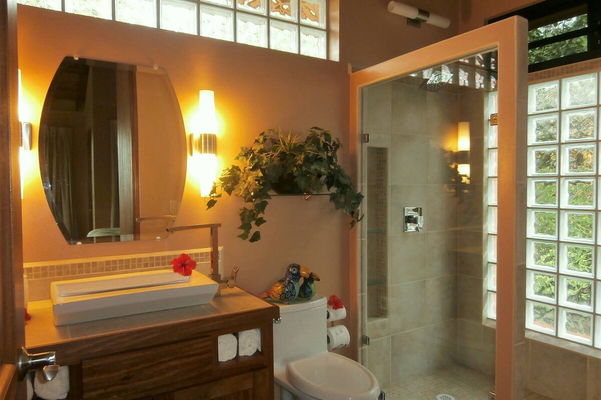 A bathroom with a wooden vanity, a white toilet, a glass shower enclosure, and a large mirror with wall sconces.