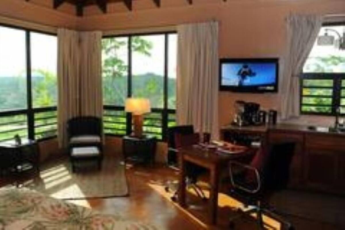 A cozy room with a wooden ceiling, a television mounted on the wall, a dining table with chairs, and large windows offering a view of greenery outside.