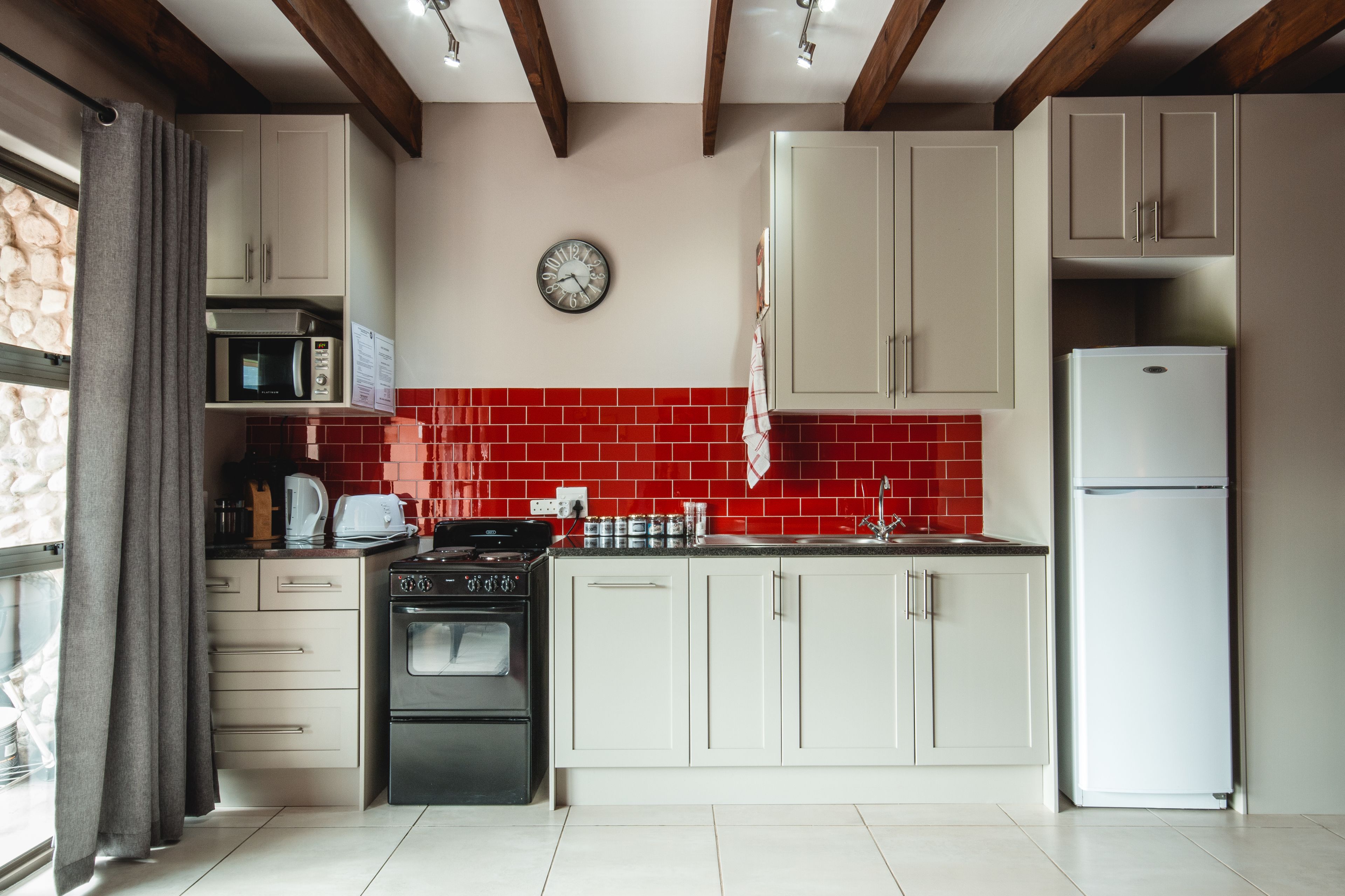 Een keuken met witte kastjes, een rode tegelwand, een magnetron, een oven, een koelkast en een klok aan de muur.